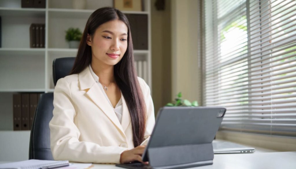 A female professional in a white blazer working on a digital tablet at a desk, illustrating a remote worker managing global earnings under the UK-US double tax treaty.