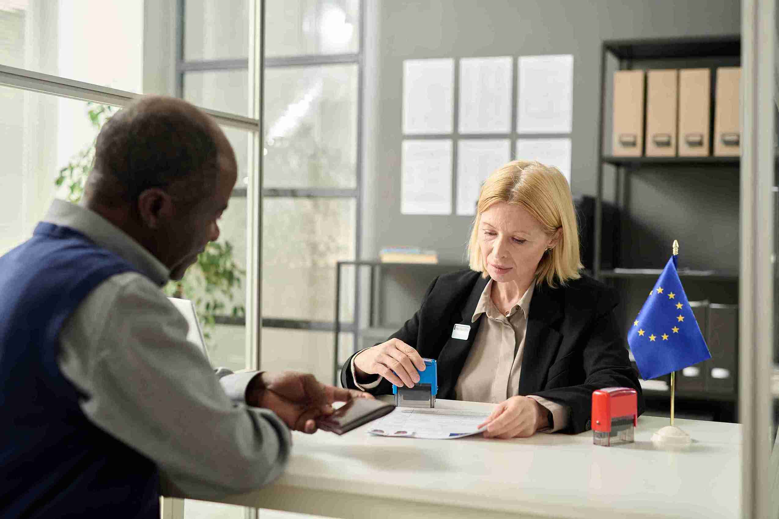 A man and woman sit at a desk, discussing with a European flag displayed prominently in the background.