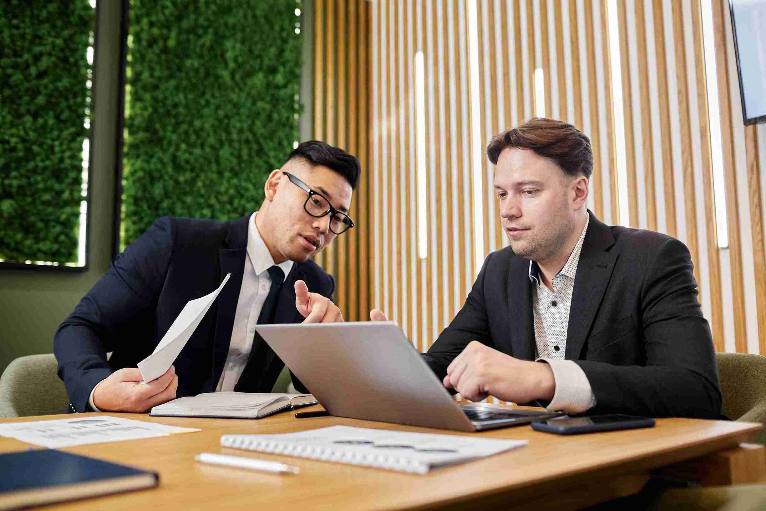 Two men in suits discussing UK Sponsor Licence details at a table with a laptop open between them.