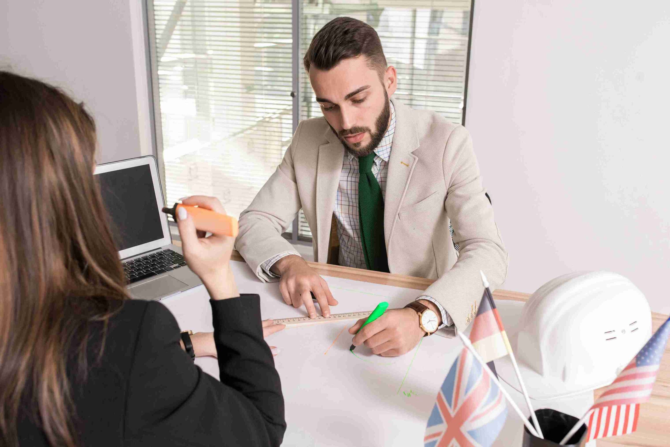 A man and woman in business attire sitting at a desk, engaged in a discussion with documents in front of them.
