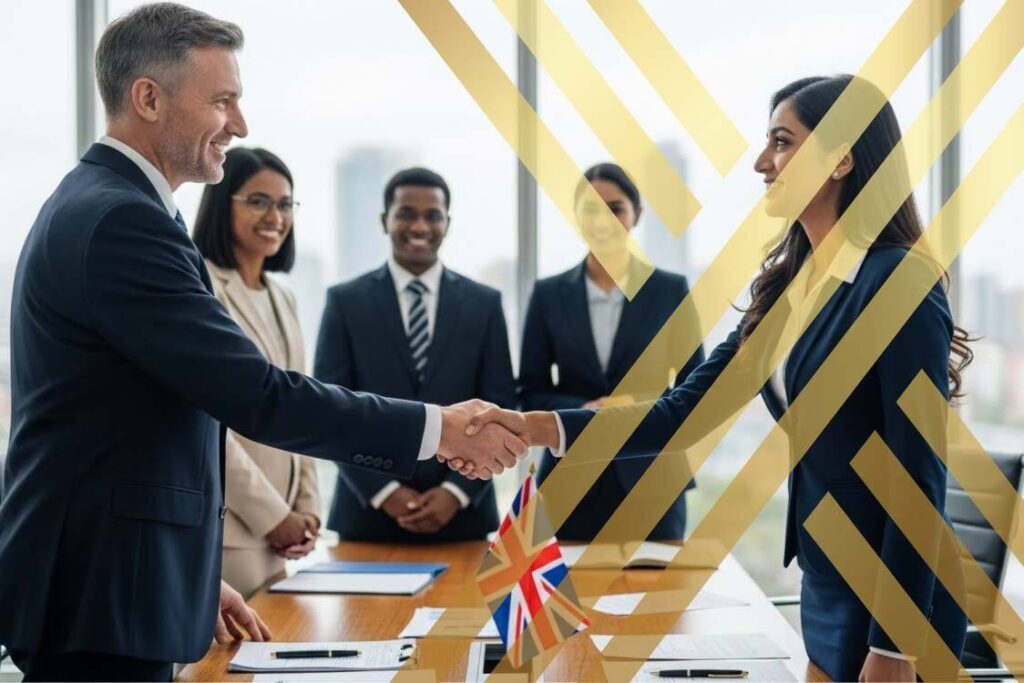 Businessperson shaking hands with an international employee in an office environment with some other officials in the picture too and some documents and a small UK flag on table.