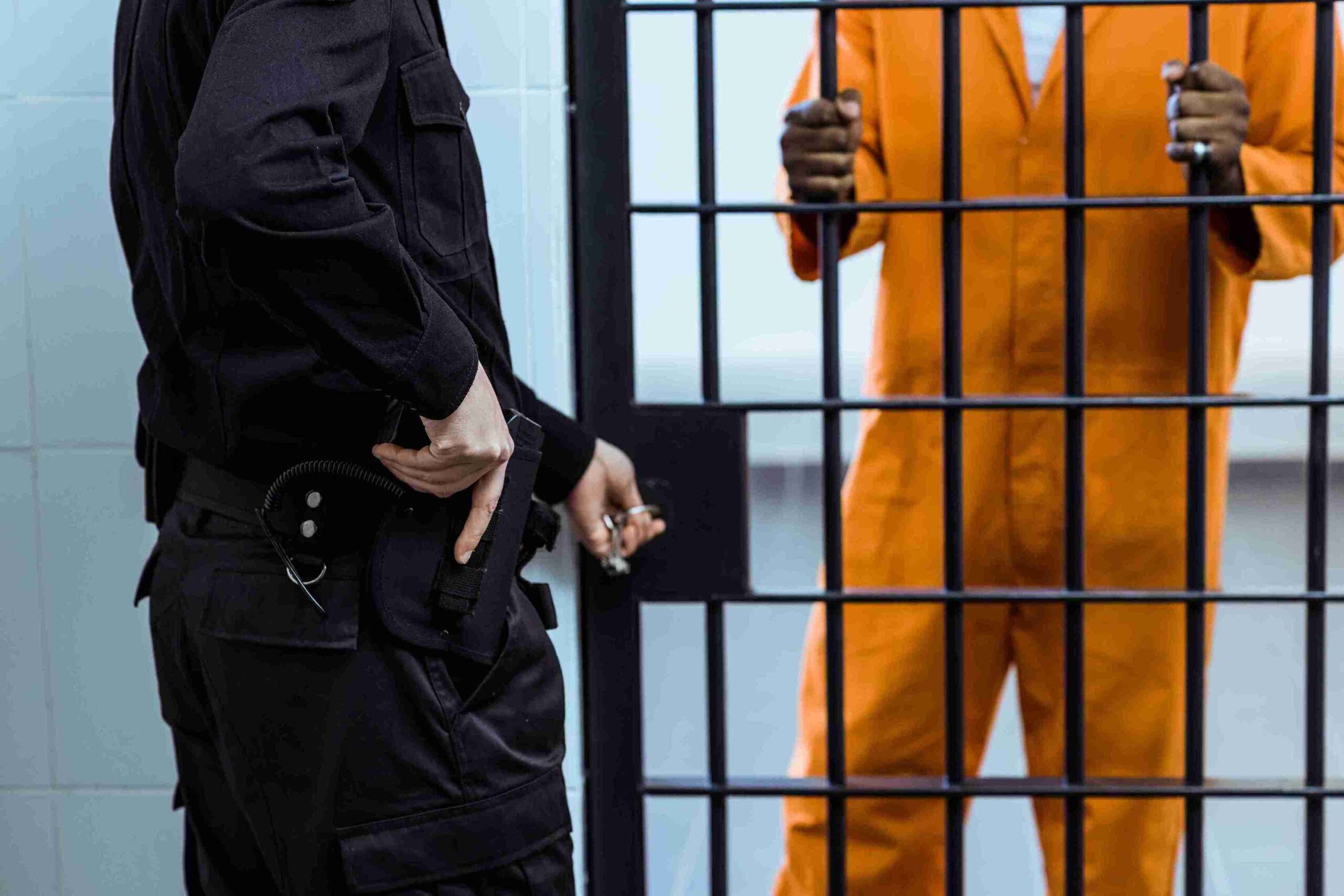 A man in orange prison clothes stands in front of a jail cell, looking solemn and reflective.