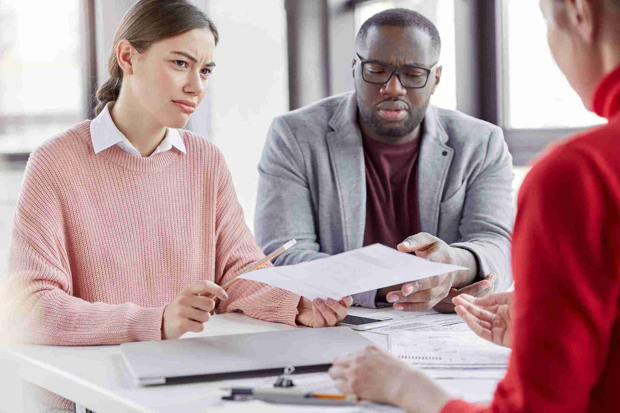 Three people at a table with papers; a woman is speaking to another while the third listens attentively.