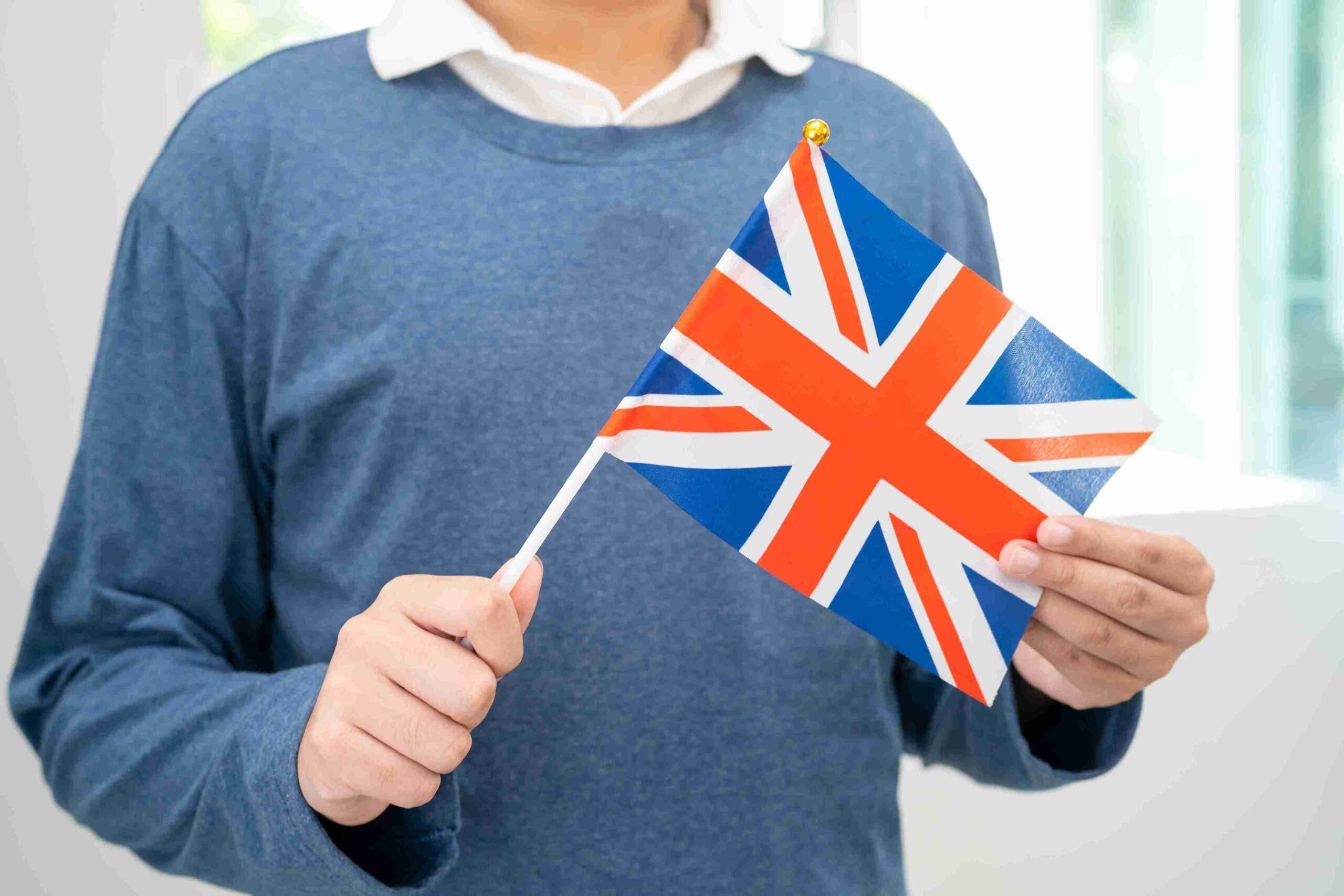 A young man proudly holds a British flag, smiling against a bright background.