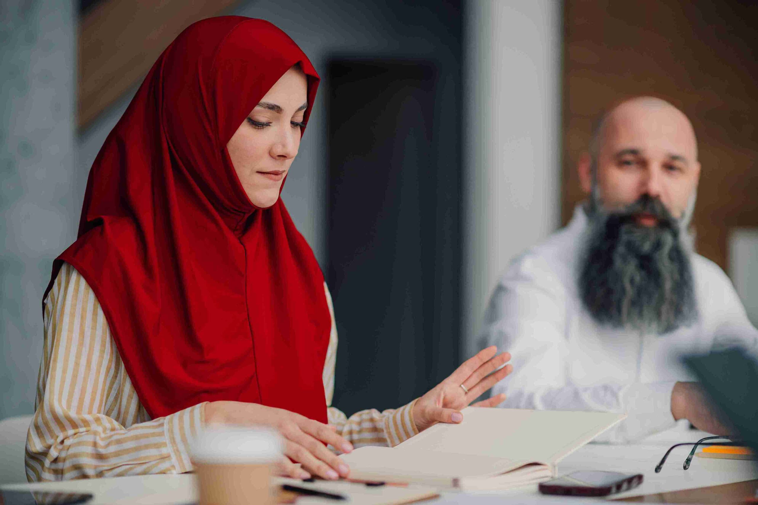 Woman in red hijab reading notebook at desk with man observing, guiding family arrangements respecting both faith and law.