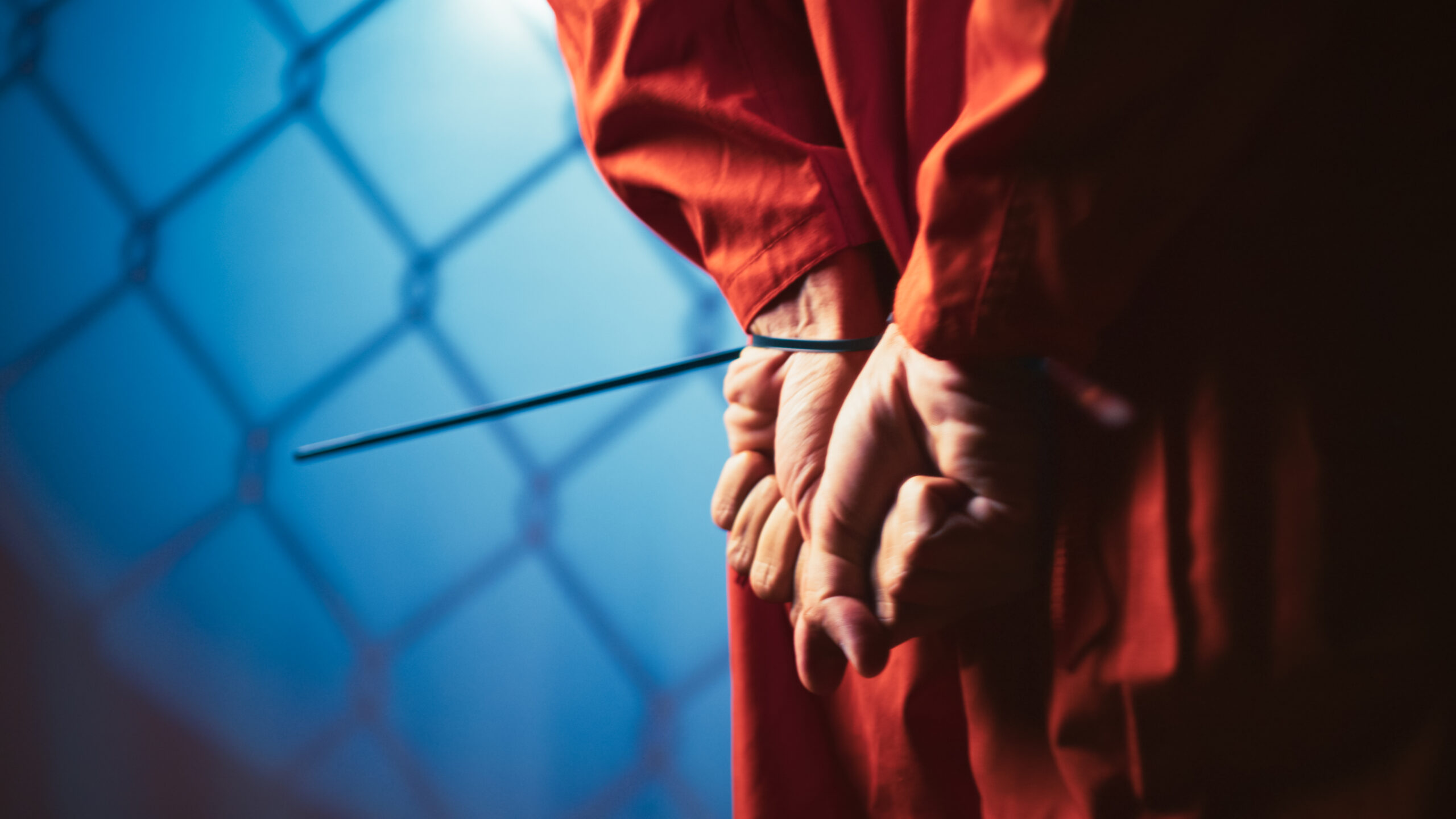 A person in an orange prison uniform stands holding a stick, looking directly at the camera.