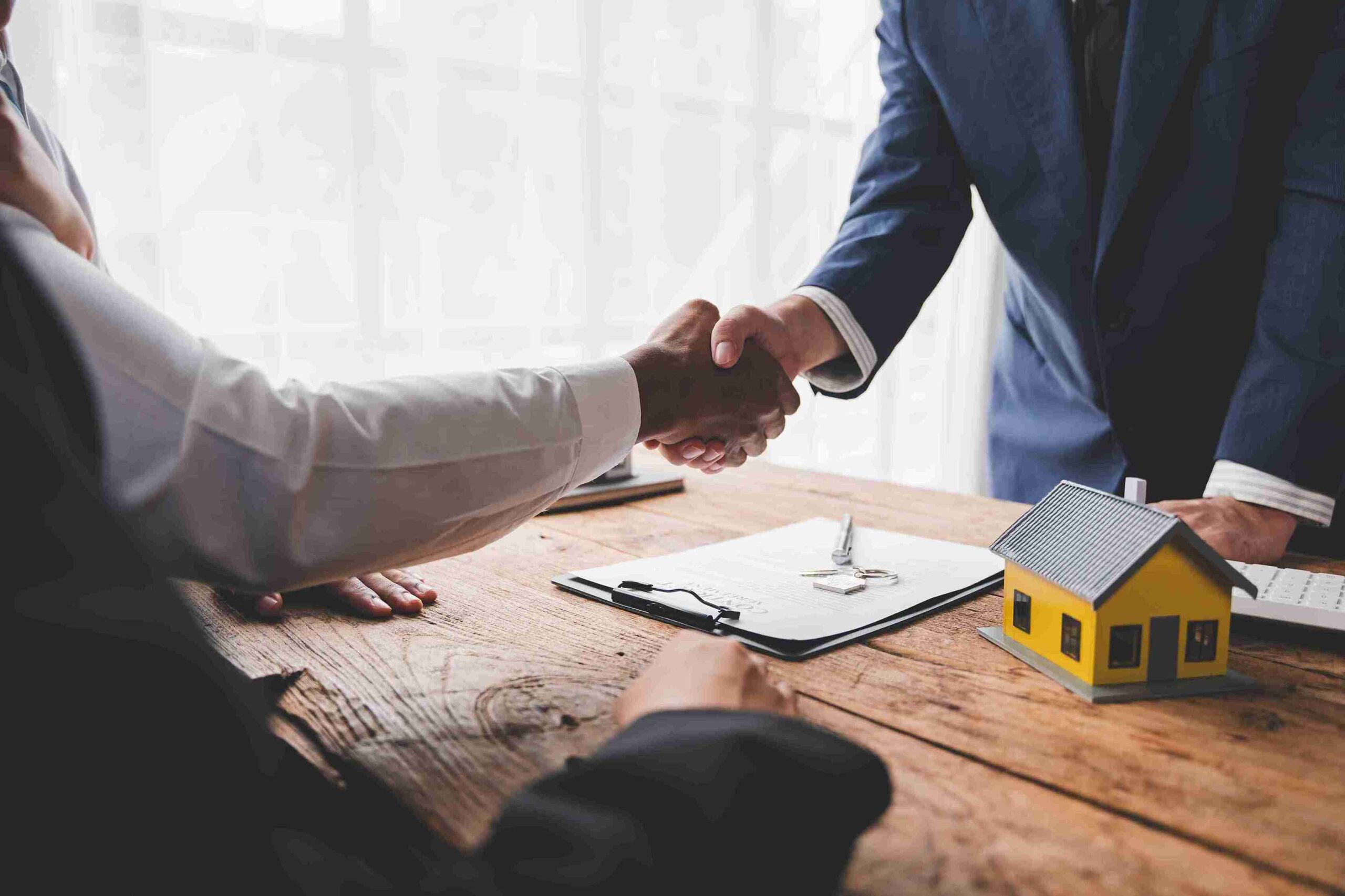 Two people shaking hands over a table with a house model, symbolizing a successful property transaction.
