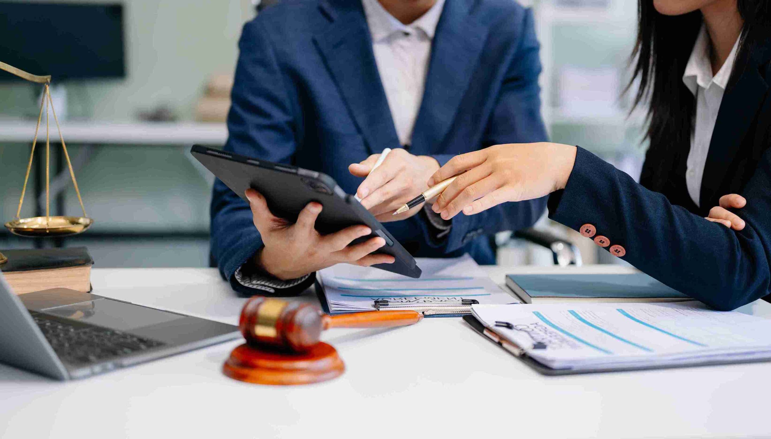 Two business professionals at a desk, one holding a tablet and the other a gavel, discussing a legal matter.