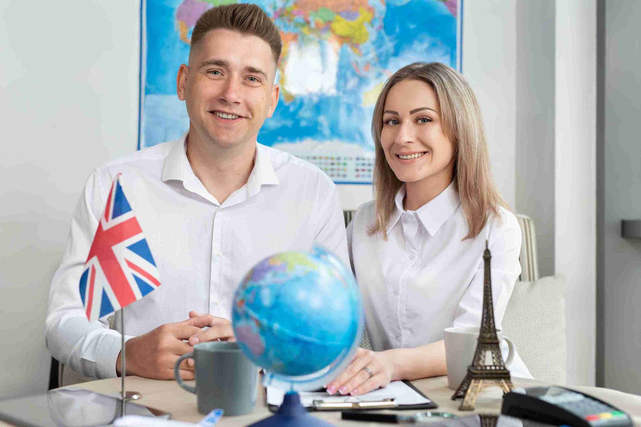 A man and woman stand by a desk featuring a globe and a British flag, engaged in conversation.